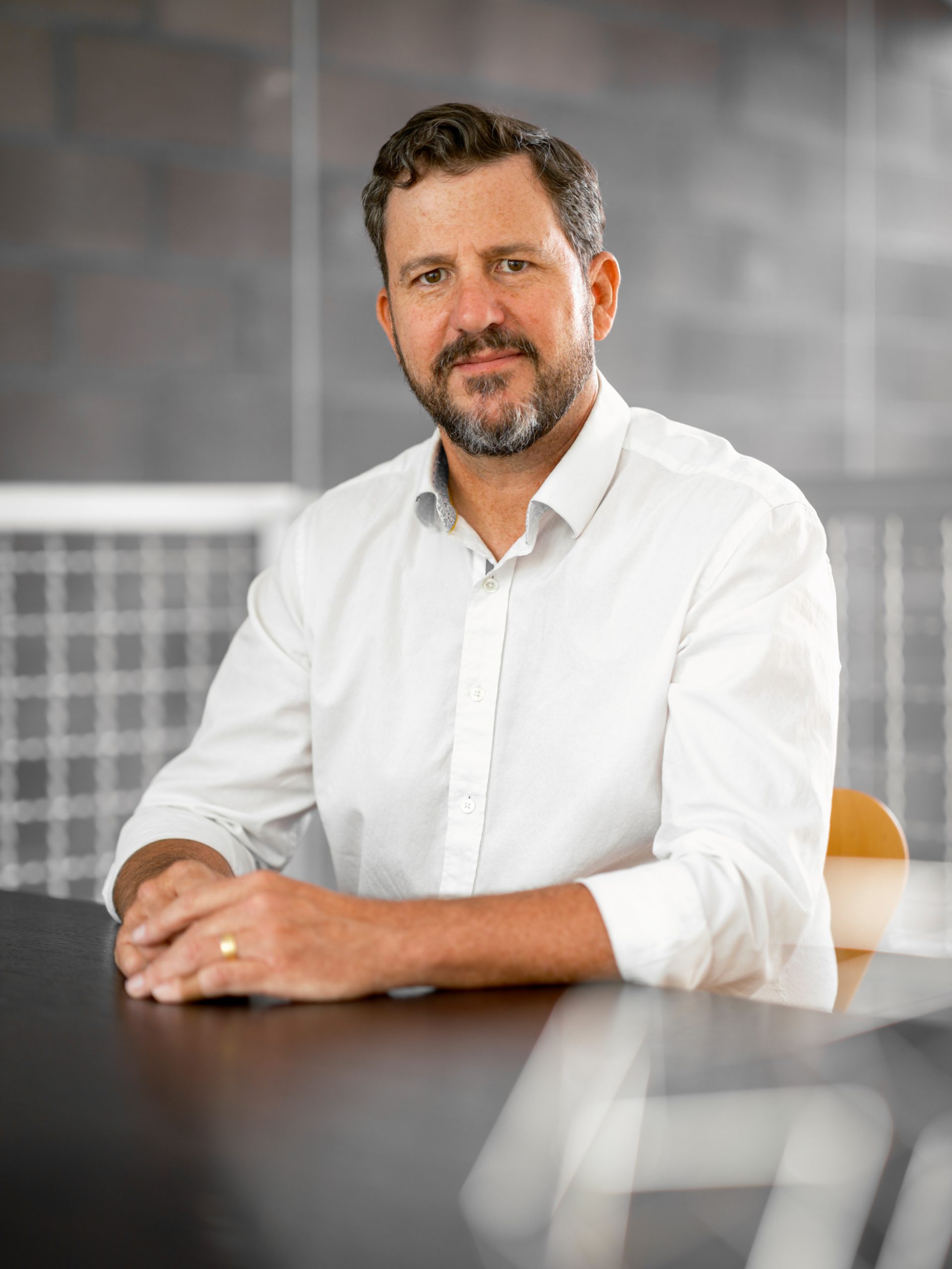 Photograph of a man with dark hair and beard, wearing a white shirt, sitting at a desk.